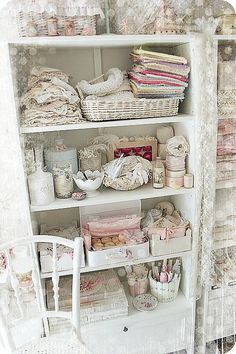 a shelf filled with lots of items next to a white chair and wall covered in lace