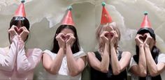 four women with hats covering their faces in front of a white wall holding hands up to their mouths