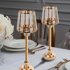 two gold candlesticks on a white table with silverware and flowers in the background