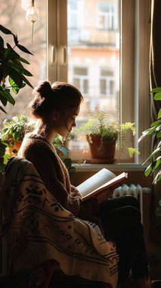 a woman sitting on a chair reading a book in front of a window with potted plants