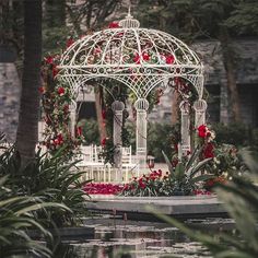 a white gazebo with red flowers and greenery on the ground next to water