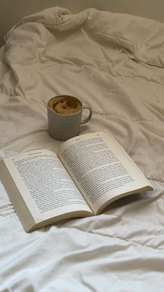 an open book sitting on top of a bed next to a cup of hot chocolate