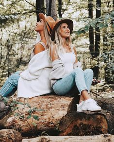 two beautiful women sitting on top of a rock in the woods wearing hats and sweaters