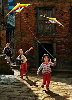 three children running with kites in the air on a brick walkway next to an old building