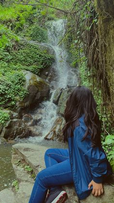 a woman sitting on top of a rock next to a small waterfall in the forest