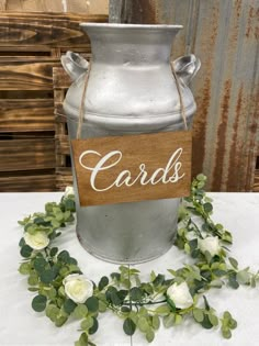 a metal jar with cards on it sitting next to some white flowers and greenery