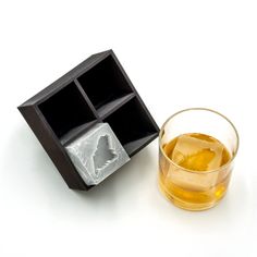 an ice cube next to a glass filled with liquid on a white surface, in front of a black box