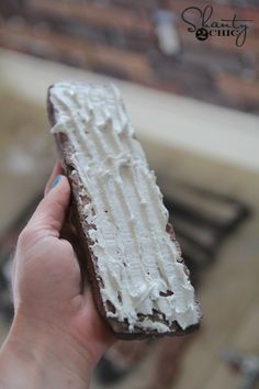 a hand holding a piece of cake with white frosting on it and a brick wall in the background