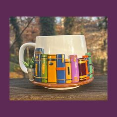 a coffee cup with colorful books on it sitting on a wooden table in front of some trees