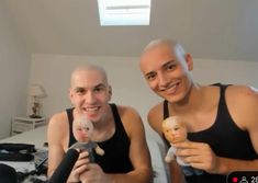 two men sitting on a bed holding dolls in front of their faces and smiling at the camera