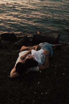 a person laying on the ground next to some water and rocks, with their arms around each other