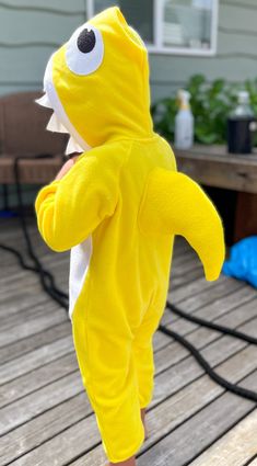 a toddler in a yellow shark onesie standing on a deck with his arms out