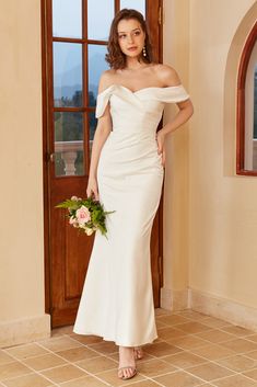 a woman standing in front of a door wearing a white dress and holding a bouquet