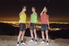 three men standing on top of a mountain holding up their glasses with the city lights in the background