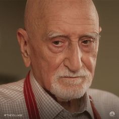 an older man with white hair wearing a gray shirt and red suspenders looking at the camera