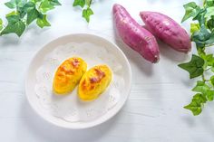 two purple sweet potatoes on a white plate next to green leaves and ivy, one is cut in half