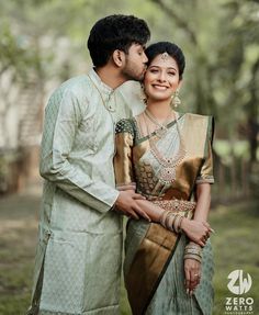 a man and woman standing next to each other in front of trees with their arms around each other