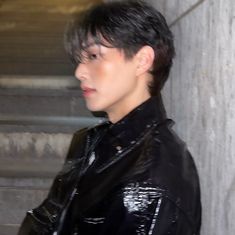 a young man in black leather jacket standing next to a cement wall and looking off into the distance