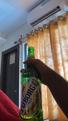 a person holding up a bottle of beer in front of a window with drapes