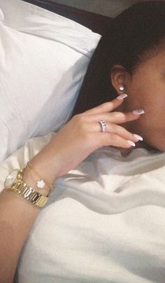 a woman laying in bed with her hand on her face and nails painted white, wearing gold bracelets