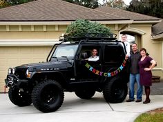 three people standing in front of a black jeep with a christmas tree on top