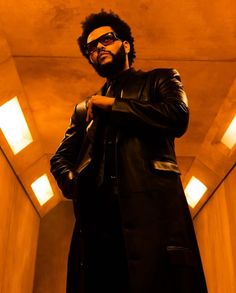 a man in a black coat and sunglasses standing in a room with lights on the ceiling