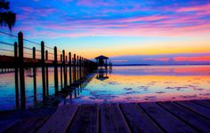 a wooden dock sitting on top of a lake under a purple and blue sky at sunset