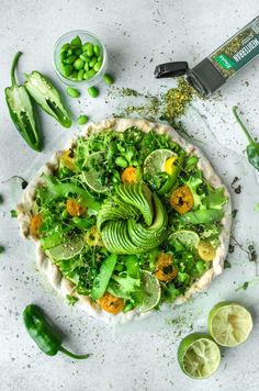 a green vegetable pizza on a white plate with limes and seasoning next to it