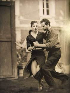 an old photo of two people dancing in front of a building, one man is holding the woman's leg