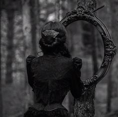 a woman is standing in the woods looking at her reflection in a mirror that hangs on a tree