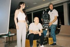 two men and a woman standing in a living room with a camera on the floor