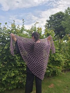 a woman wearing a purple crocheted shawl standing in front of some bushes