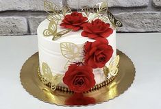 a white and gold cake with red roses on top, sitting on a table next to a brick wall