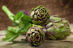 three artichokes with green leaves and purple flowers on a wooden table top