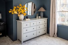 a white dresser sitting next to a window with yellow flowers on top and a mirror above it