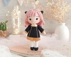 a crocheted doll with pink hair and black dress stands on a table next to flowers