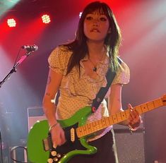 a woman holding a green guitar in front of a microphone and some lights on the stage