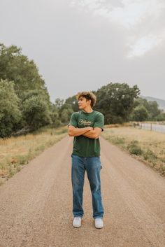 a man standing in the middle of a dirt road with his arms crossed and looking off to the side