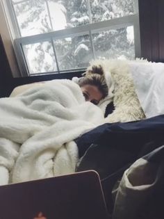 a woman laying in bed under a blanket next to a window with snow on it