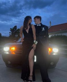 a man and woman in formal wear standing next to a car at night with the lights on