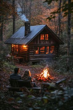 two people sitting around a campfire in front of a log cabin with a lit fire