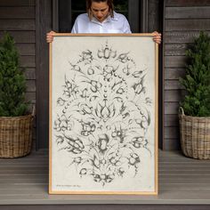 a woman holding up a framed artwork with flowers on it in front of some plants