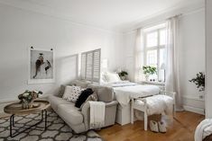 a living room with white walls and wood flooring is furnished with an ottoman, chair, coffee table, bed, and rug