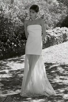 black and white photograph of woman in dress standing on path with bushes behind her, looking off to the side