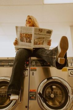 a woman is sitting on top of a washing machine and reading the paper while looking at her phone