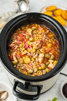 a crock pot filled with chicken and vegetable soup next to silver spoons on a white table