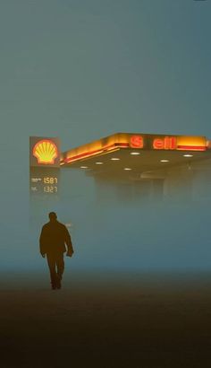 a man walking past a shell gas station in the foggy night with neon lights