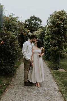 a man and woman standing next to each other in front of some bushes on a path