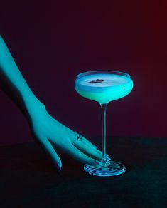 a woman's hand next to a cocktail glass with blue liquid in it on a table