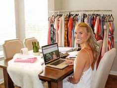 a woman sitting at a desk with a laptop computer in front of her and clothes on racks behind her